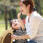 晴れた公園で、白い服を着た女性が幼い女の子を抱きしめ、2人で笑っている様子