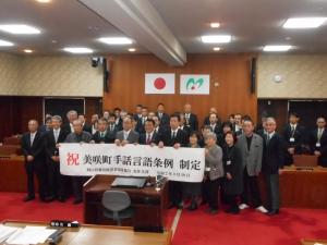 スーツ姿の人たちが「祝 美崎町 手話言語条例 制定」と書かれた横断幕を持っている集合写真
