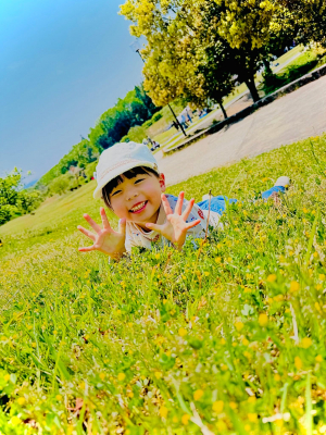 子どもが芝生の上に肘をついた形でうつ伏せになり、両手をパーの状態にして笑顔でカメラ目線で写っている写真