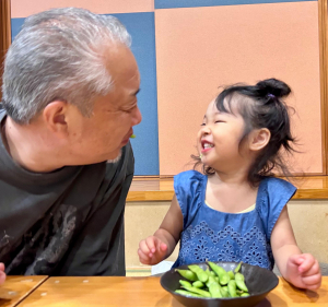 青い服を着た女の子の前に皿の上に乗った枝豆が沢山あり、女の子の横に高齢の男性がいて女の子と見つめ合っている写真