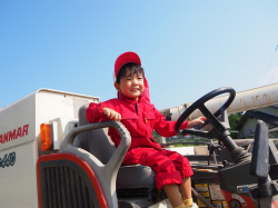 赤いつなぎを着た男の子がトラクターの助手席に乗りハンドルを手で触っている写真