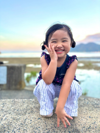 女の子が海を背景にしゃがみながら片手を頬に当てながらにっこり笑っている写真