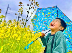 緑色の服を着て青い傘を持った男の子が黄色い花が咲くお花畑の横で笑っている写真