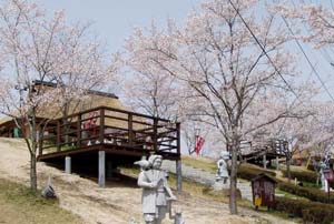 斜面に桜の花が咲き、傍らに石造が立っている民話の村の写真