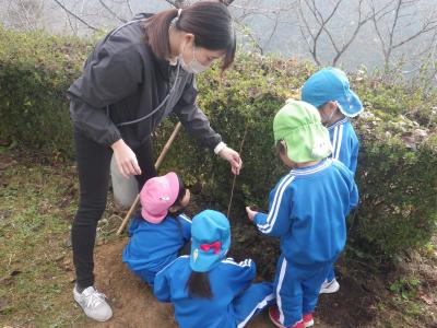 マスクをした女性が園児たちに説明しながら苗木の根元へ土を被せている写真