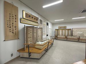 展示室の壁に掛け軸や額装作品が掛けられており、その前に展示ケースが並んで配置されている写真