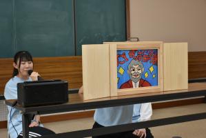 机の上で紙芝居を上演し、横でマイクを持った女性が読み聞かせをしている写真