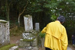 森の中の古い石碑の前で、黄色い上着を着た人物が古い石碑を指し示している写真