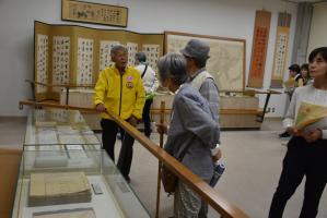 展示室で、黄色い上着を着た人が、ガラスケースの前で来館者に資料について説明している写真