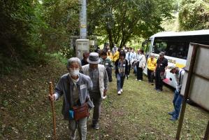 観光バスから森林の中へ続く道を杖をついた白髪の女性を先頭に歩き出している人々の写真