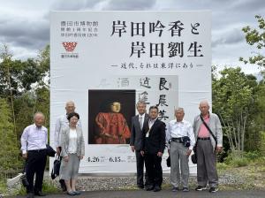 岸田吟香と岸田劉生と書かれた大きな展示会看板の前で、スーツを着た7人の人が並んで記念撮影している写真