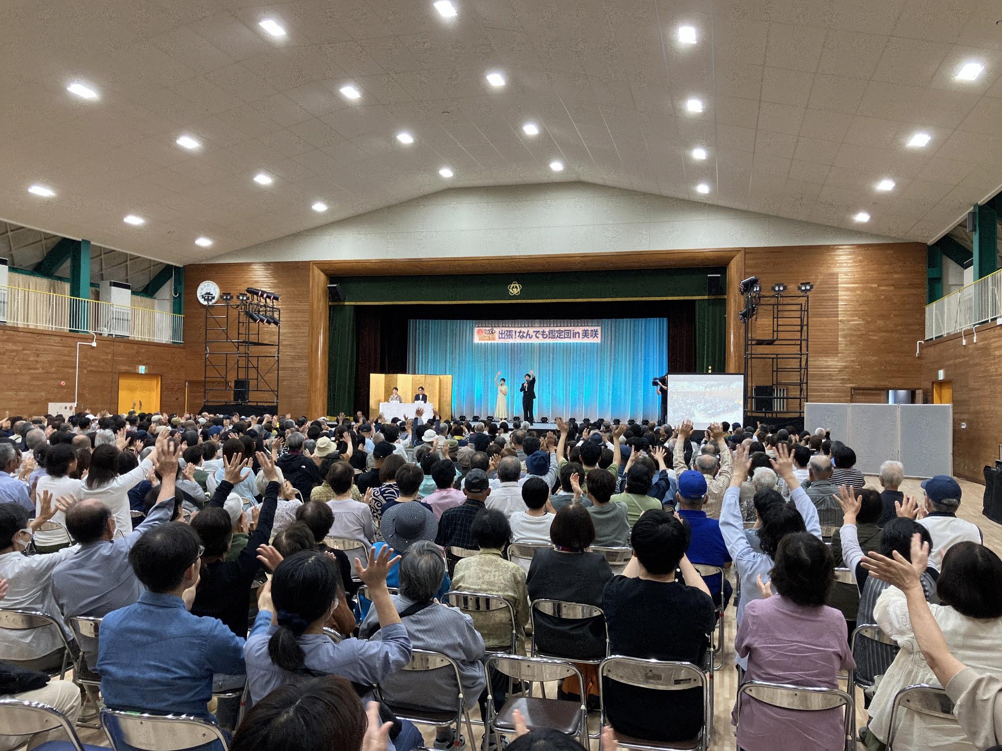 「なんでも鑑定団in美咲」と書かれた横断幕がステージ上にあり、ステージの上の二人が手を挙げており、会場の人々のうち何人かも手を挙げている写真