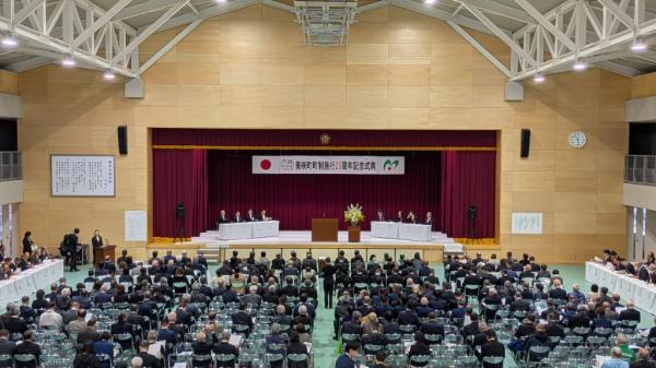 体育館のような会場の奥にステージが見え、手前に客席があり人々が集まっている写真