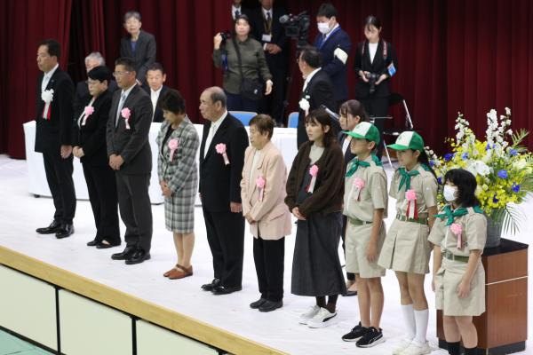 胸元にピンクの花とリボンを付けた人々がステージ上に横一列に並んでいる写真