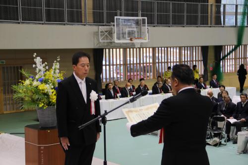 賞状を両手で持つスーツ姿の男性と、賞状を目の前にして起立しているスーツ姿の男性の写真