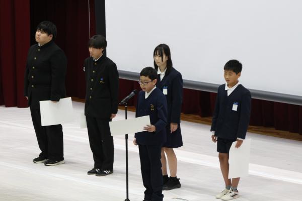 奥に手に香美を持った四人の生徒がいて、手前で一人の生徒が紙を見ながらマイクに向かって話している写真