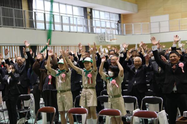 ベージュのつなぎを着た児童3名を中心に両手を挙げている客席の人々の写真