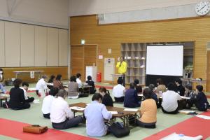 テーブルごとにグループを作って座りながら司会者の話を聞いている大人たちの写真