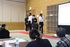 マイクを使ってスクリーンに映っている内容を伝えている学生と聴衆の方々の写真