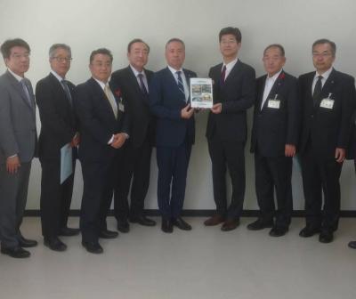 一緒に書類を持ってこちらに見せる2人の男性と、その横に並び立つスーツ姿の人々の写真