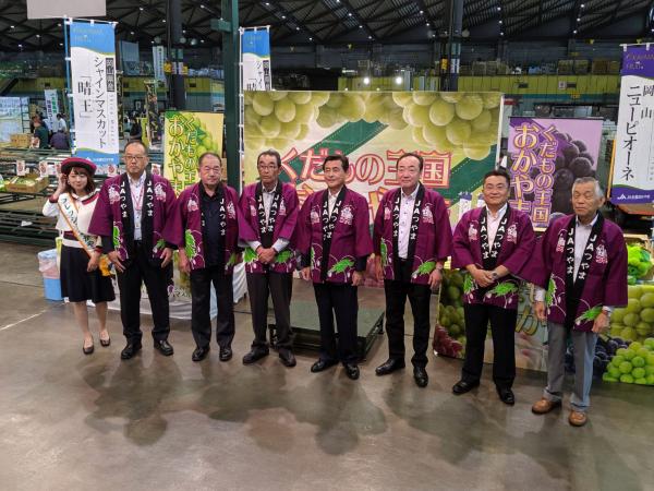 ぶどうのポップやのぼりがたくさん設置された売り場の前で紫色の法被を着た男性達が並んでいる写真