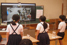 3人の児童が部屋の壁側に設置されたモニターを見ながら立っている写真