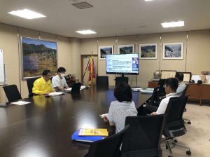 会議室で大きなテーブルを囲んで着席し、前方に設置されたモニターを見る人々の写真