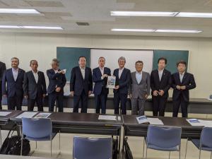 書類を手に持つ2人の男性とその横に並ぶ男性達の写真