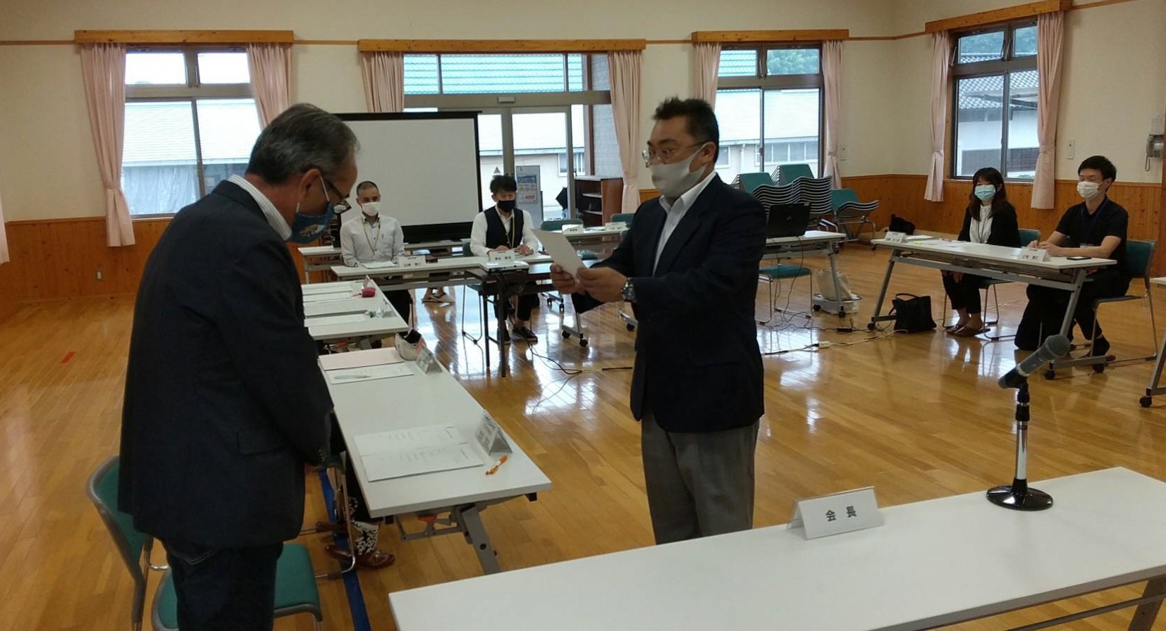 長机の前に立ち、男性に書類を手渡そうとする男性とそれを着席して見守る数人の人達の写真