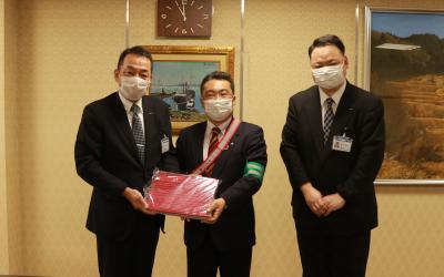 赤い長方形のものを手に持つ2人の男性と、1人のスーツ姿の男性達の記念写真