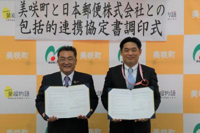 バックパネルを背に、書類をこちらに開いて見せて持ち笑顔を見せる、二人のスーツ姿の男性の写真