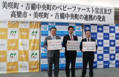 バックパネルを背に、文字が書かれた白い厚紙を胸の前に掲げている3人の男性達の写真