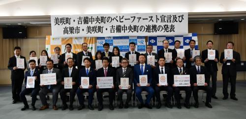 前後二列に並び、一人一人が書類を胸の前に掲げ持つスーツ姿の男女の集合写真