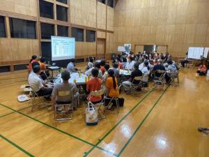 体育館に設置されたプロジェクター画面の前で多数の参加者が話し合っている定期共有会の写真
