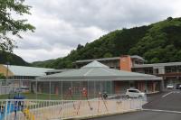 手前にフェンスで囲まれた遊具があり、奥に多角形屋根の建物が見える「みさキッズ あさひ」の外観写真