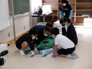 毛布を使ってカエル型の人形を搬送しようとする児童たちの写真